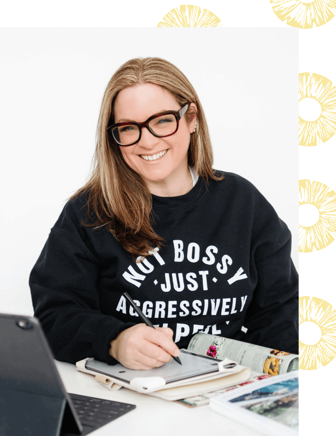 Top Ajax REALTOR® Laura McBride smiling while wearing a “Not Bossy, Just Aggressively Helpful” sweatshirt, working with a tablet and magazines at her desk