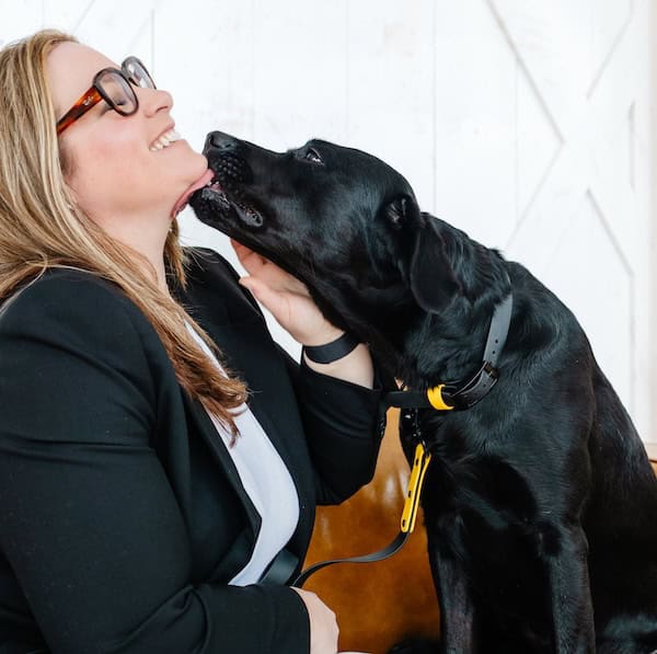 Laura McBride smiling while her black Labrador, Ripp, licks her face affectionately