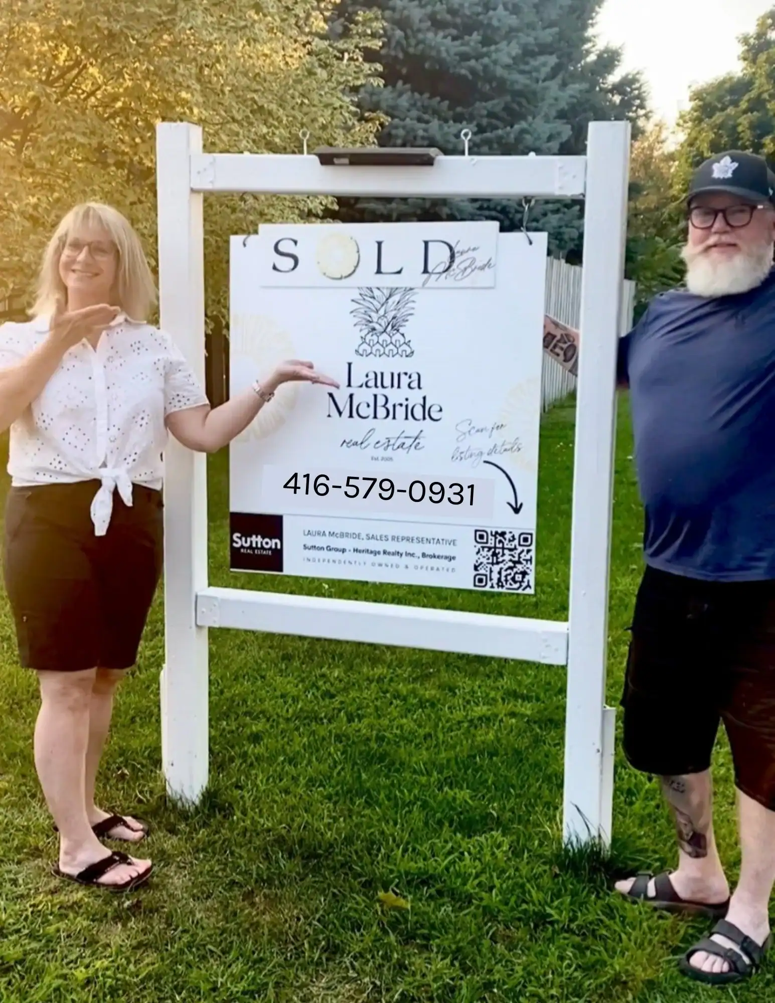 Two happy home sellers standing in front of a Laura McBride Real Estate sold sign, celebrating that their sale will fund a Junior Green Thumbs classroom garden