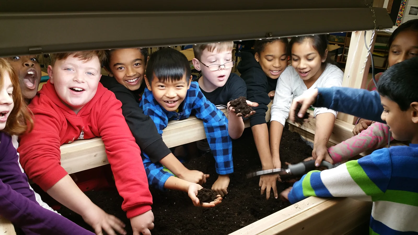 Students from the first Junior Green Thumbs pilot class smiling and reaching into a raised garden bed filled with fresh soil during their first planting activity