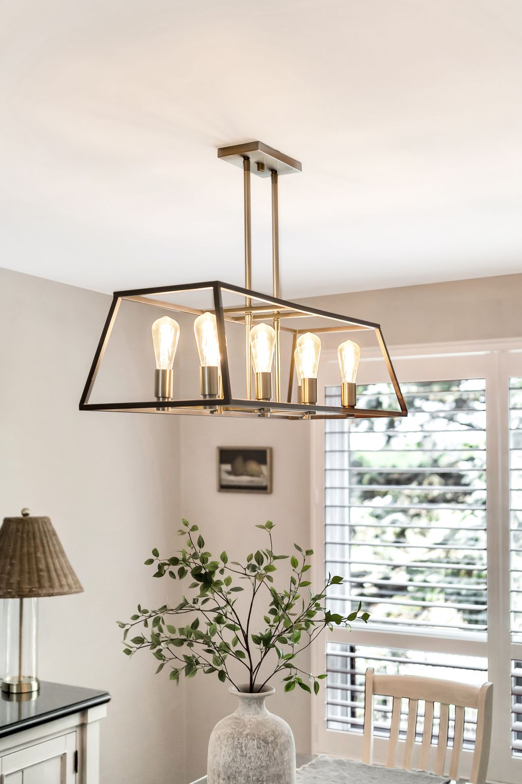 Modern rectangular pendant chandelier with exposed Edison-style bulbs above a dining table with greenery.