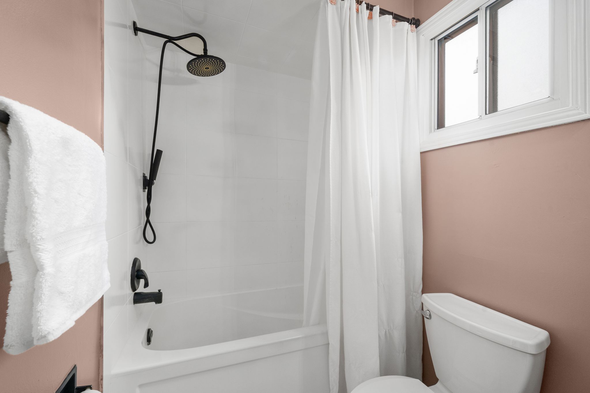 Full view of renovated bathroom with white tub, matte black rain shower, white curtain, and dusty rose walls