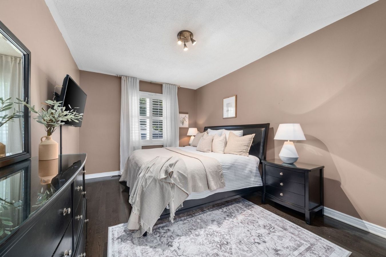 Primary bedroom with hardwood floors, large window with California shutters, neutral bedding, and black furniture.