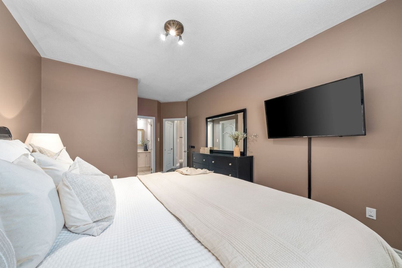 Primary Bedroom with Ensuite View: Spacious primary bedroom showing ensuite entry, dresser, wall-mounted TV, and soft neutral décor.