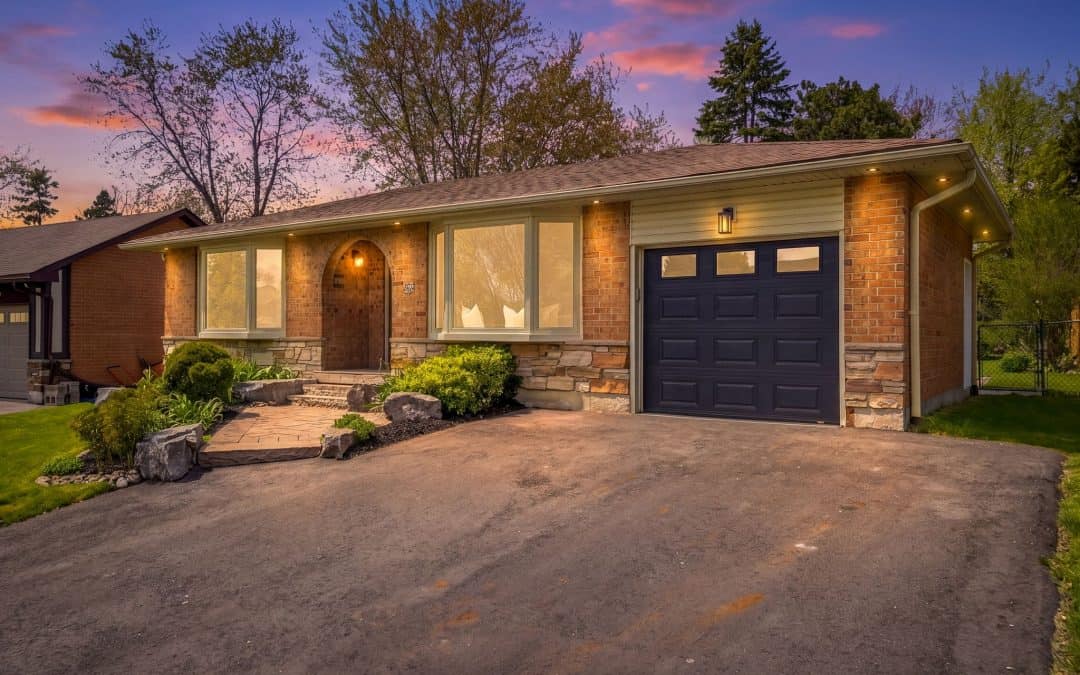 Front exterior of a brick Ajax bungalow for sale at dusk, with landscaped gardens, a dark garage door, wide driveway, and mature trees.