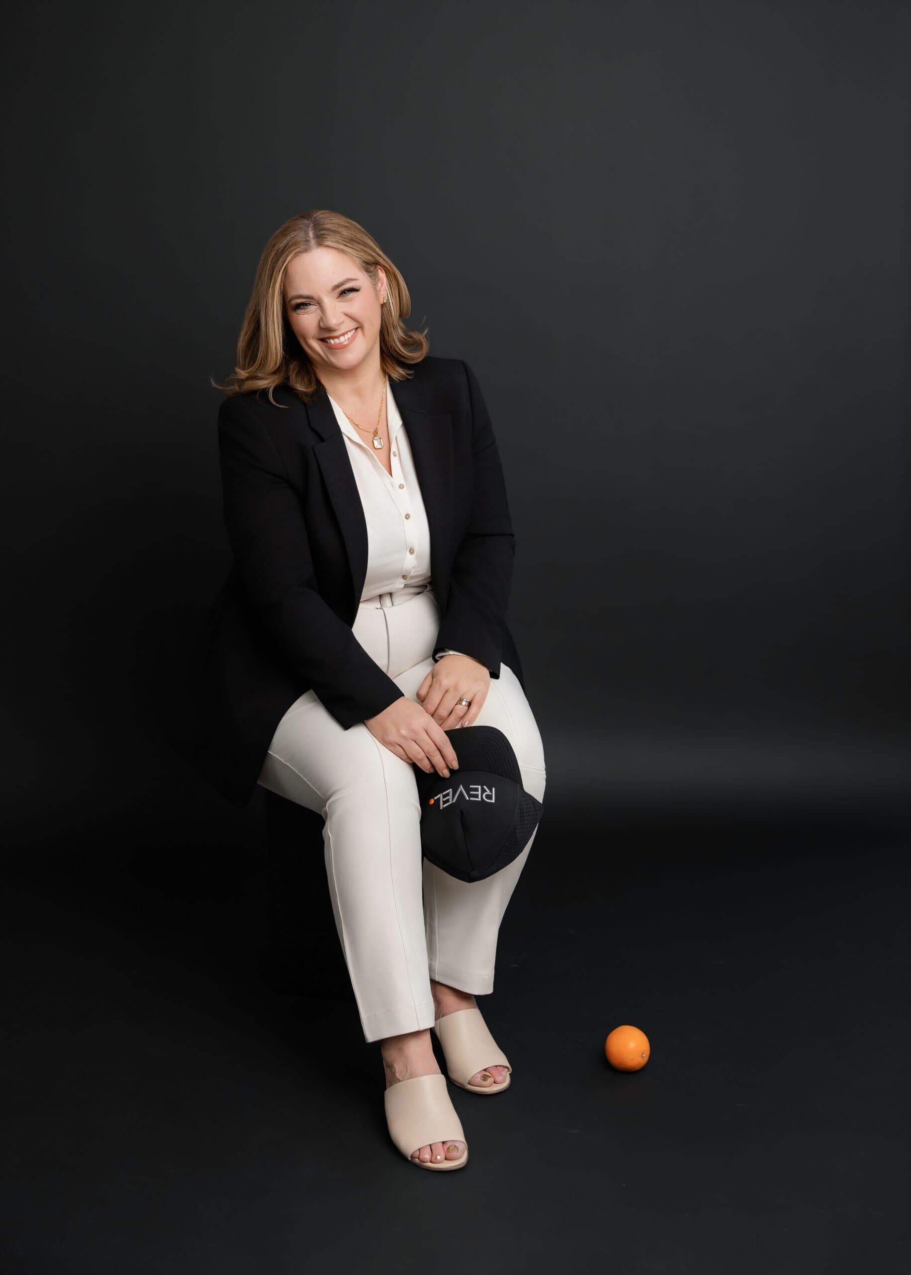 Laura McBride, Ajax Realtor, seated in a black blazer and white outfit against a dark studio backdrop, holding a REVEL Realty hat.
