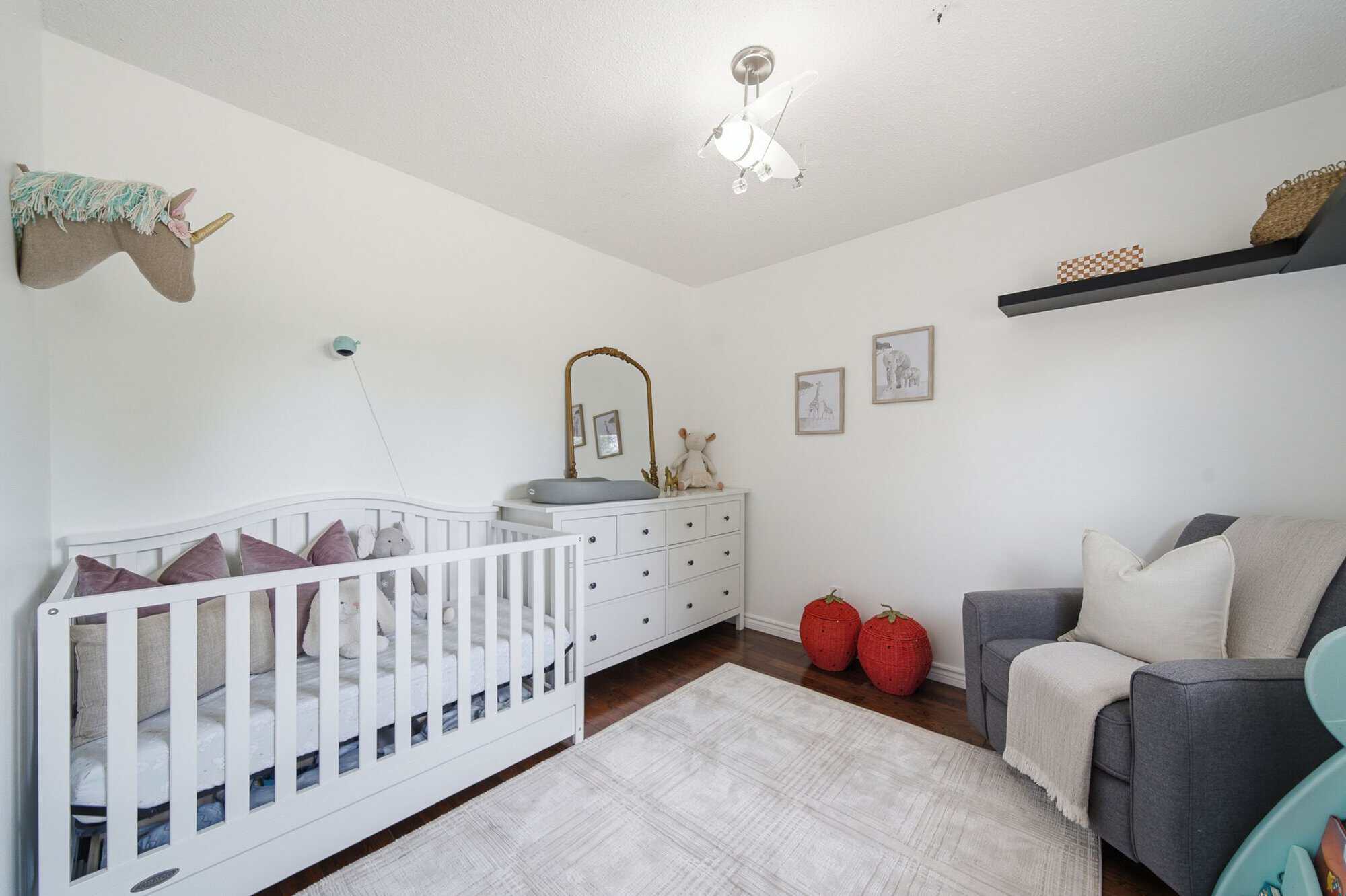 Bright nursery with a white crib, dresser, grey armchair, hardwood floors and soft neutral décor.