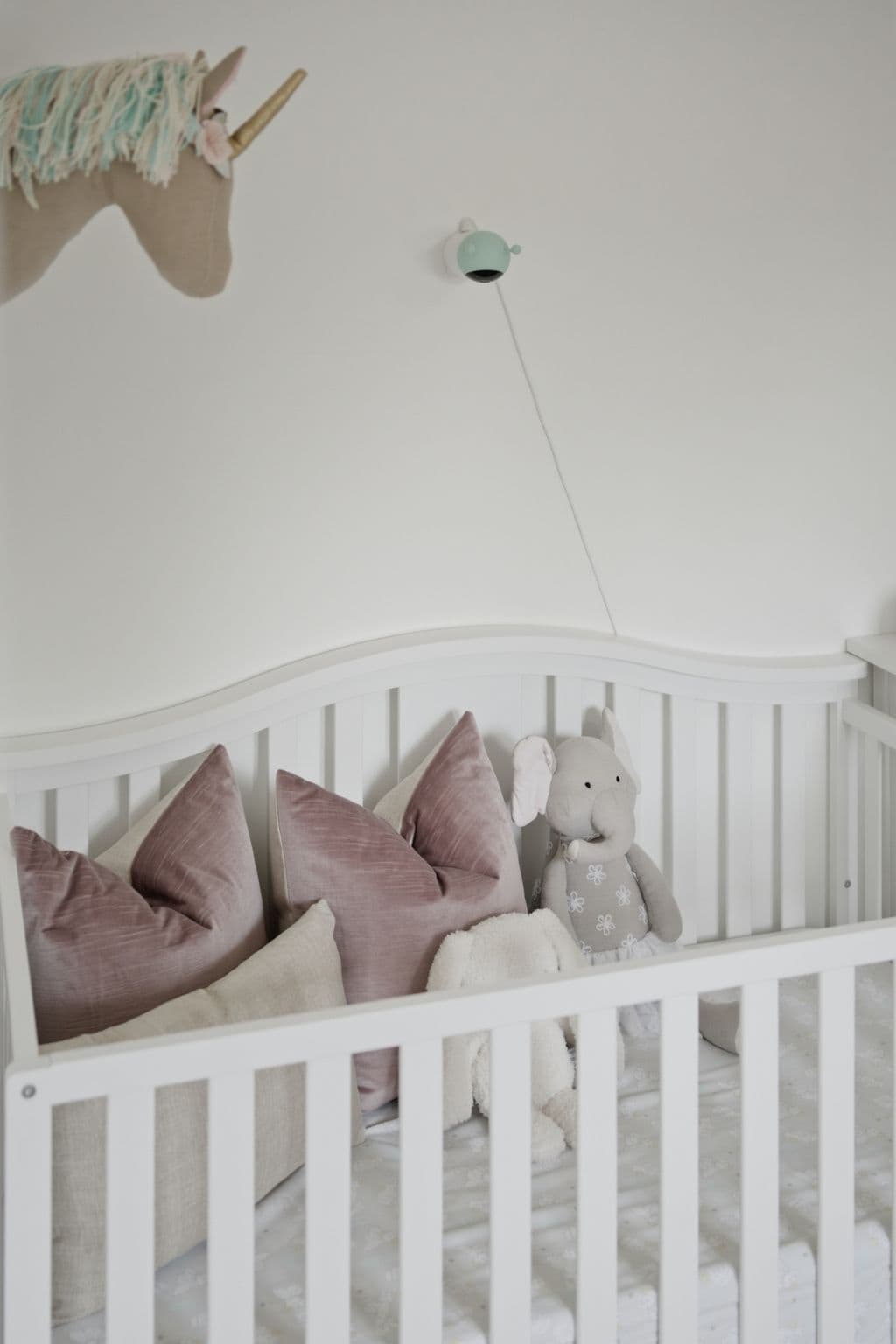 Close-up of a white crib styled with soft pillows, plush toys and a decorative unicorn wall hanging.