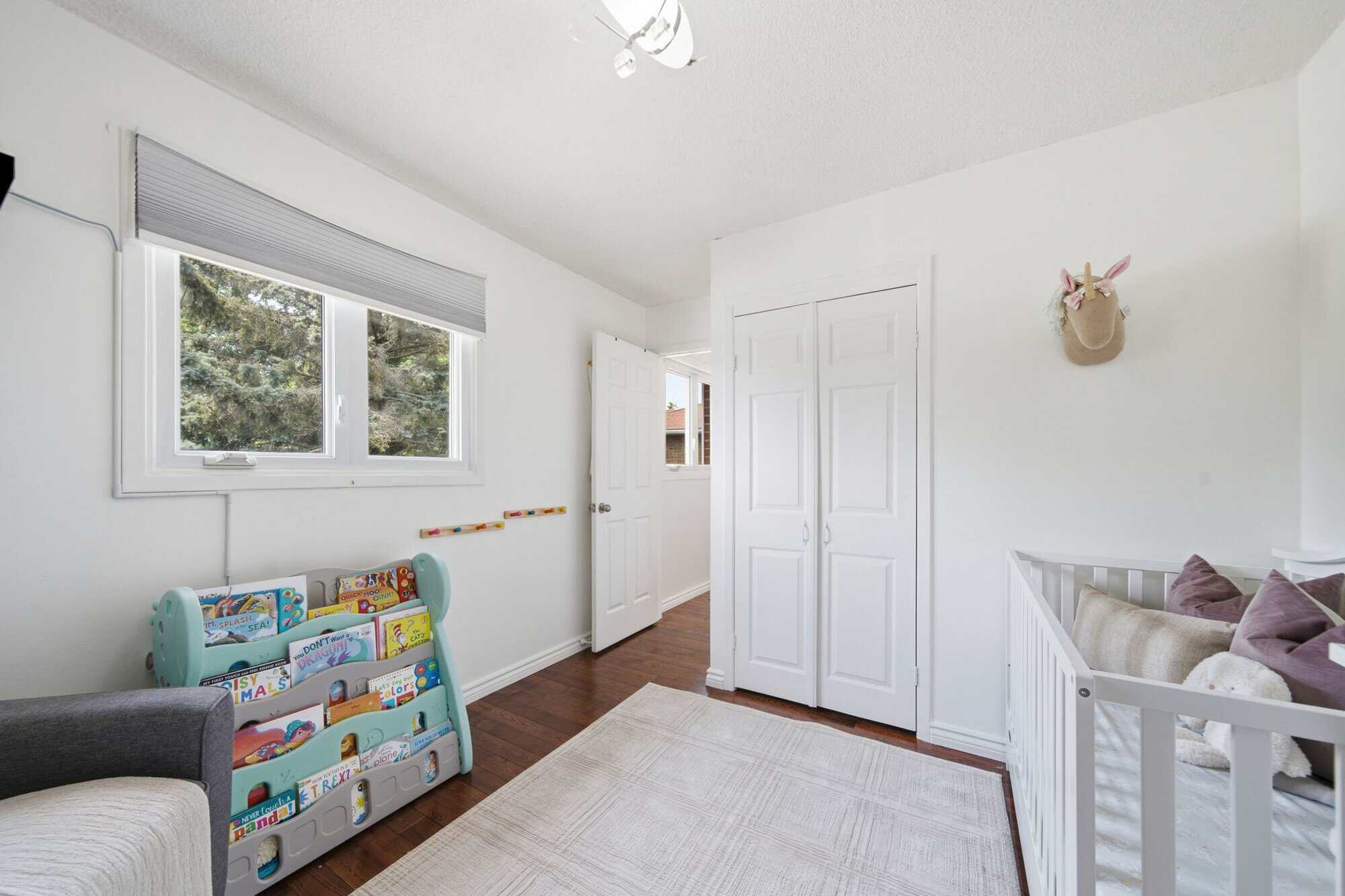Nursery with a large window, white closet, turquoise book rack, hardwood floors and part of a white crib.