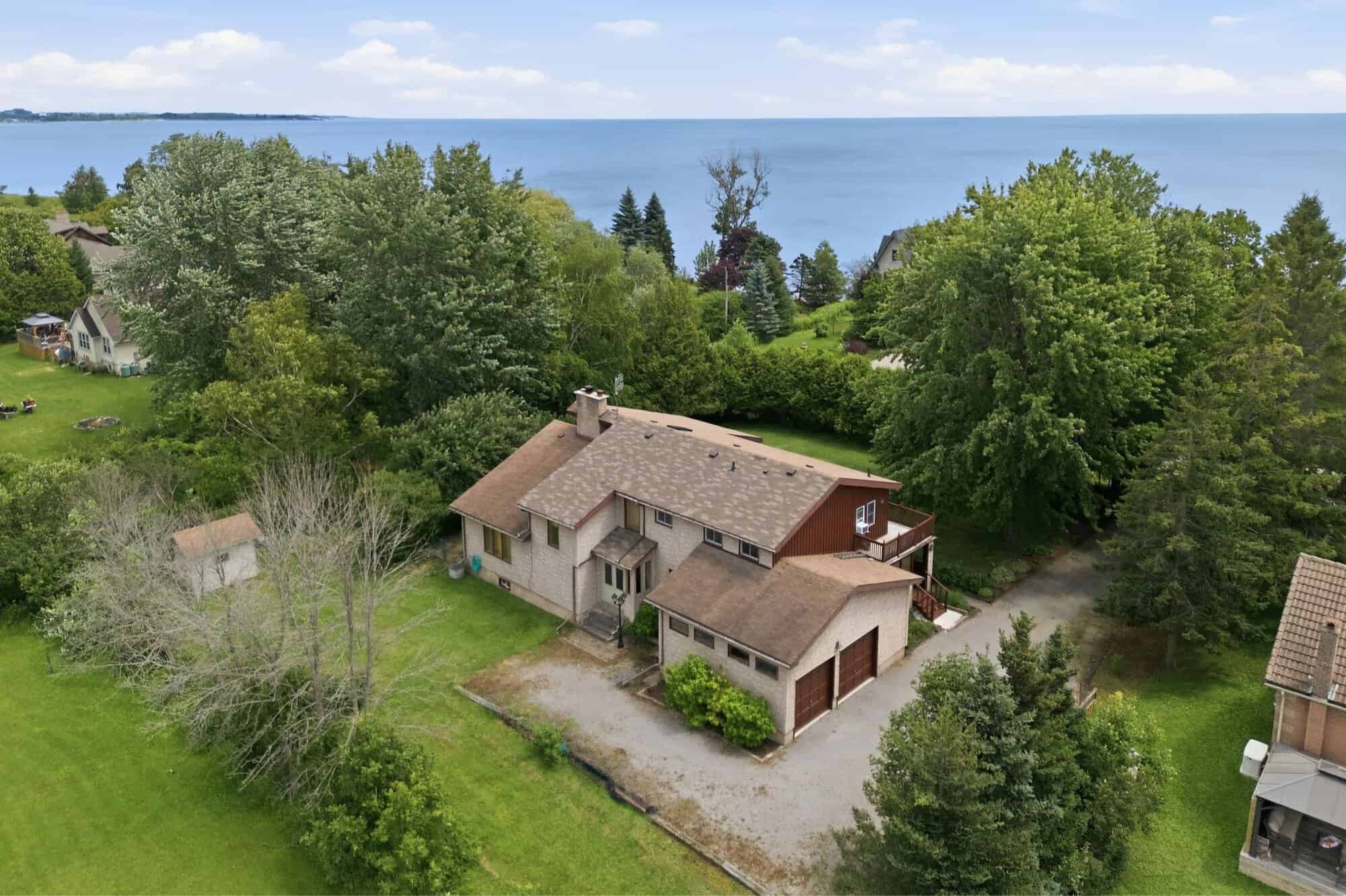 Aerial view of a substantial custom stone home with an attached oversized garage, mature trees, and glimpses of Lake Ontario beyond.