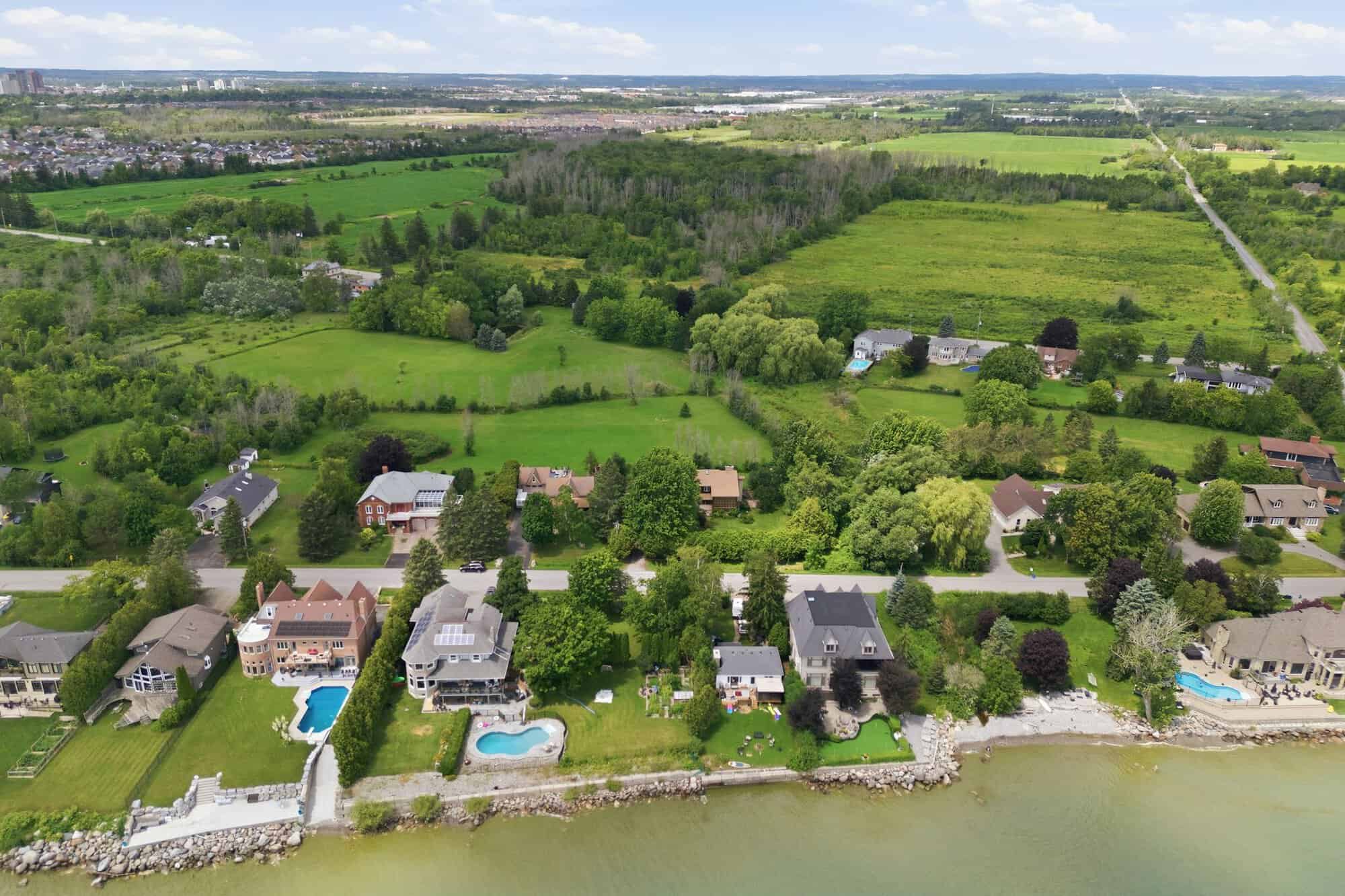 Aerial view looking inland from the Lake Ontario shoreline, showcasing luxury waterfront homes, expansive estate lots, mature trees, and the surrounding greenbelt landscape of South Ajax.