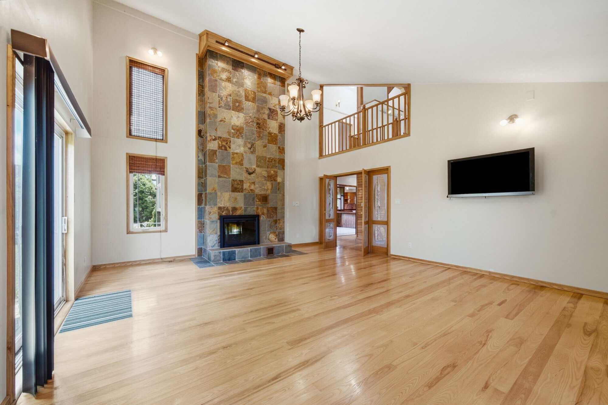 Spacious great room with vaulted ceilings, hardwood floors, oversized windows, and a dramatic floor-to-ceiling double-sided stone fireplace.